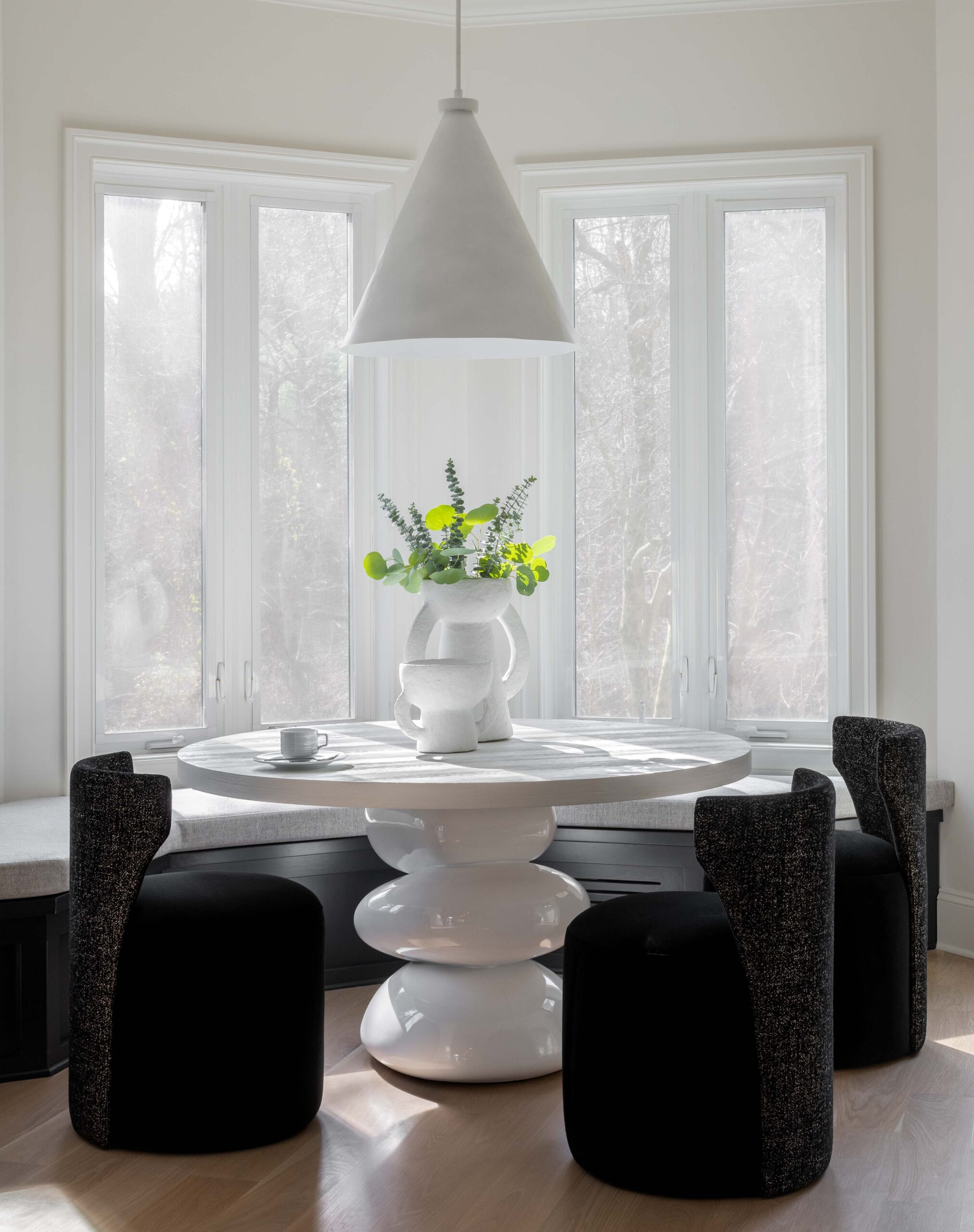 A white circular table surrounded by dark chairs in front of large windows spilling in natural light